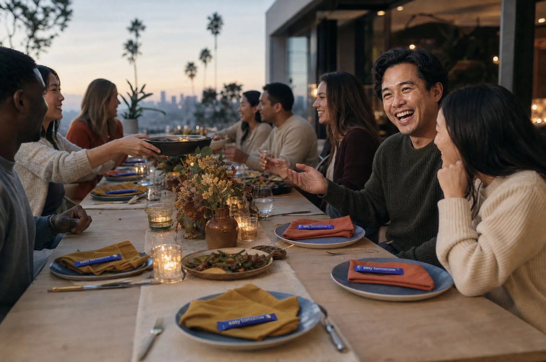 Friends laughing around a long Friendsgiving table at dusk, an Easy Tomorrow stick on every napkin
